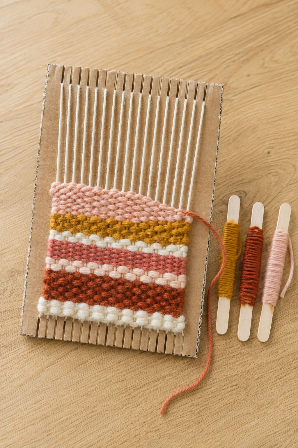 Small notched cardboard loom wrapped with colorful yarn strands and a mini wooden weaving shuttle stick